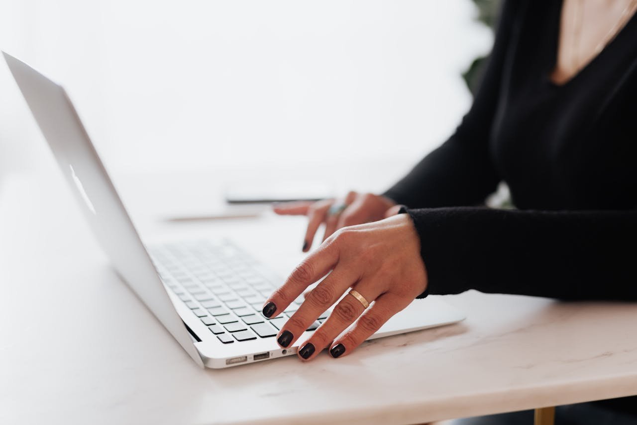 Webmaster with dark nail polish types on a silver laptop, which is placed on a white table.